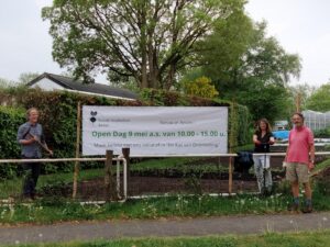 drie mensen staan bij een banner waarop de open dag van de stichting sociale voedseltuin op 9 mei 2026 wordt aangekondigd. De banner staat bij een moestuin en een kas